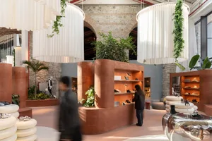 Modern shoe store interior featuring curved terracotta display walls, large white cylindrical fabric pendant lights with hanging greenery, exposed brick arches, and shoppers browsing sneakers.
