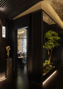 Modern restaurant entrance featuring vertical dark wood slats, an indoor tree garden with uplighting, a scholar rock sculpture on a marble pedestal, and a view into the dining area.