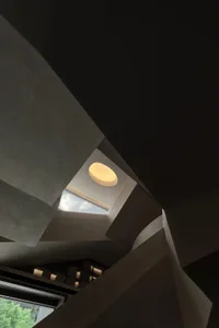 Low-angle view looking up at a sculptural concrete staircase featuring sharp geometric angles, a large circular skylight, and warm recessed lighting.