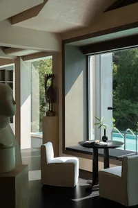 Modern minimalist reading nook featuring beige armchairs, dark round table, wood paneling, abstract sculptures, and large windows overlooking a swimming pool and greenery.