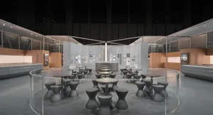 Modern exhibition space with rows of dark concrete hourglass-shaped stools and round tables inside a large circular glass partition, set in an industrial-style hall with concrete floors and wooden display walls.