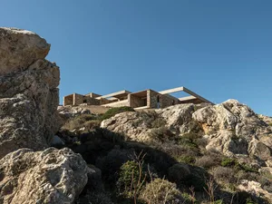 Modern stone and concrete house built into a rocky cliffside with dramatic overhangs and open terraces, surrounded by Mediterranean scrub vegetation under a clear blue sky.