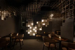 Modern restaurant interior featuring a large geometric light installation made of illuminated cubes, dark textured plaster walls, and warm brown leather dining chairs arranged around round wooden tables.