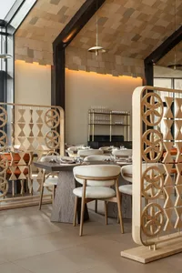 Modern Japanese-inspired restaurant interior featuring light wood dining tables, cream upholstered chairs, and ornate wooden room dividers with floral and geometric patterns under a wooden shingle ceiling.