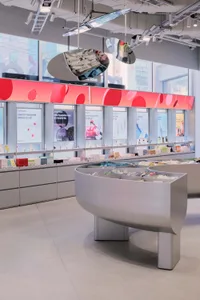 Modern cosmetics store interior featuring a large curved grey display island, red accent wall with circular patterns, product testing stations, and large windows with city views.