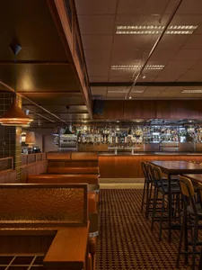 Interior view of a classic pub featuring wooden booths with textured leather upholstery, dark wood bar counter, patterned carpet, and warm copper pendant lights.
