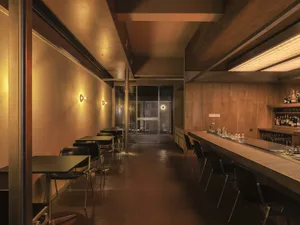 Interior view of a narrow modern bar with warm wood paneling, black bar stools, pendant lighting, and a view toward glass doors at the far end.