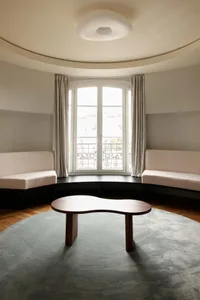 Modern minimalist living room featuring a curved bay window with built-in upholstered benches, a free-form walnut coffee table, and a large round blue-gray rug under a circular ceiling light.