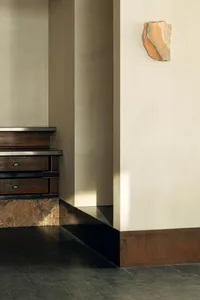 Modern minimalist interior corner featuring a dark corten steel staircase base, dark tiled floor, neutral plaster walls, and an abstract orange stone sculpture mounted on the wall.