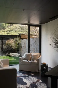 Modern living room interior featuring a cream armchair, dark wood ceiling, stacked stone wall, and large glass doors opening to a green garden with a patterned area rug in the foreground.