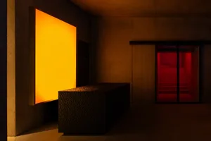 Brutalist concrete interior featuring a textured concrete bench, a glowing warm orange light panel on the left wall, and a red illuminated staircase visible through glass doors on the right.