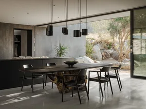 Modern dining room featuring a massive live-edge wood dining table, black leather chairs, concrete walls, and large glass doors opening to a rocky desert garden.
