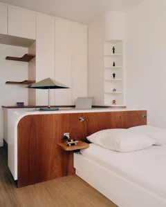 Modern minimalist bedroom with a curved walnut wood headboard, white built-in cabinetry, floating wooden shelves, and a corner display niche with figurines.