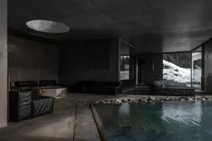 Modern brutalist indoor swimming pool with black leather modular sofas, marble tables, and large glass windows revealing a snowy mountain forest outside.