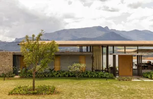 Modern single-story mountain house featuring concrete walls, vertical wood slats, large glass windows, and lush tropical landscaping with a dramatic mountain range in the background.