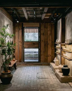 鄉村風陶藝工作室室內，回收木牆面、裸露天花板木樑、石材地磚、大型盆栽，以及展示陶瓷作品的嵌入式層架。