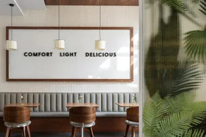 Modern restaurant dining area with grey upholstered booth, round walnut wood chairs, wooden tables, white tiled wall with large wooden frame sign reading 'COMFORT LIGHT DELICIOUS', and three pendant lights.