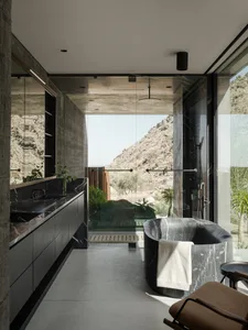 Modern luxury bathroom featuring a black marble freestanding bathtub, dark vanity with integrated sink, concrete walls, and expansive glass walls opening to a desert mountain view.