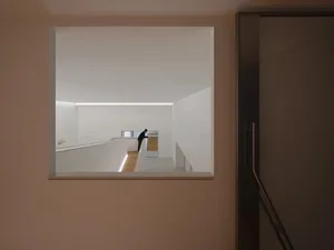 View through a square wall opening into a minimalist white hallway featuring a modern staircase with integrated LED lighting and a person standing at the far end.