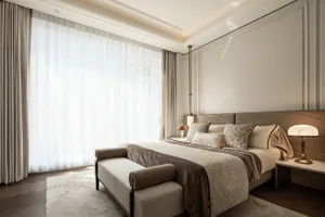Modern luxury bedroom featuring a sculptural taupe wave headboard, neutral bedding with patterned accent pillows, a pendant light, marble table lamp, and large sheer curtains allowing bright daylight.