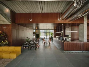 Modern industrial-style commercial cafe lounge with dark walnut wood walls, polished concrete floors, exposed stainless steel ducts, a bright yellow curved sofa in the foreground, patterned armchairs, and a long service counter under large windows.