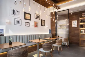 Modern cafe interior with grey upholstered banquette seating, light walnut dining tables and chairs, multiple hanging Edison bulb pendant lights, and a gallery wall of framed art.
