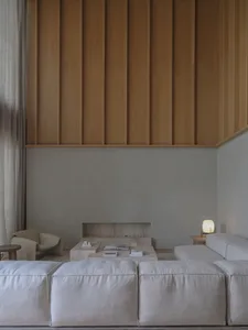Modern minimalist living room featuring a large modular beige sofa, travertine coffee tables, a built-in stone hearth, and a tall oak wood slatted accent wall under soft natural light.