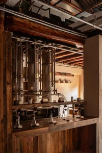 Interior view of an industrial-style distillery bar featuring large copper distillation columns, heavy reclaimed wood beams and counters, exposed metal pipes, and laboratory glassware under warm lighting.