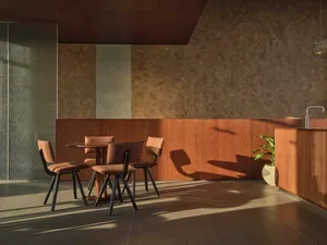 Modern minimalist dining area featuring a round walnut table, four terracotta upholstered chairs with black metal frames, wood-paneled walls, and dramatic natural light creating long shadows on a dark tiled floor.