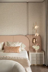 Modern luxury bedroom with blush pink curved upholstered headboard, textured beige wallpaper, white satin bedding, rose gold fur pillow, and elegant gold pendant light beside a marble nightstand with fresh flowers.