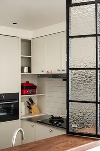 Modern minimalist white kitchen featuring handleless matte cabinets, black induction cooktop, open shelving with red multi-cooker and knife block, and a textured glass partition on the right.