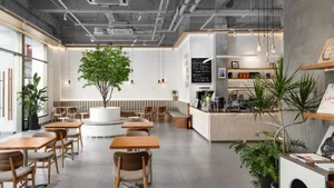 Modern industrial-style cafe interior with a large potted tree in the center, wooden tables and chairs, concrete walls, and pendant lighting.