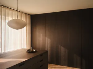 Modern minimalist bedroom corner featuring dark walnut built-in wardrobe, matching floating console with decorative tray, large rice-paper pendant lamp, and sheer linen curtains allowing soft natural light.