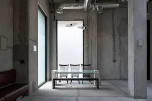 Industrial-style dining area featuring raw concrete walls, a glass dining table with black spindle-back chairs, a black bench, and two modern pendant lights suspended from an exposed ceiling with visible ductwork.