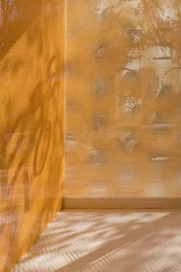 Close-up view of a golden perforated metal screen in a corner, with sunlight creating striped and dotted shadow patterns on the light-colored floor.