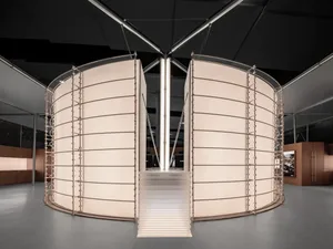 Modern art installation with two large curved fabric screens in off-white with metal frames, a central staircase leading upward, set in a spacious minimalist gallery with dark ceiling and concrete floor.