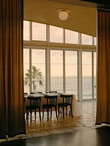 Elegant dining table set for guests in a room with large ocean-view windows, stone flooring, and heavy golden curtains at sunset.