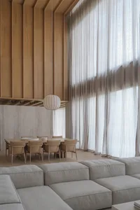 Modern minimalist dining area featuring a large oak table with beige upholstered chairs, a spherical ribbed pendant lamp, vertical oak wall paneling, and a textured concrete accent wall, viewed from a light-colored modular sofa in the foreground.