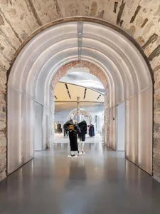 Long view down a retail corridor featuring massive layered metallic arches set within ancient stone walls, with two fashion mannequins in black designer clothing standing in the center.