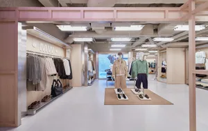 Modern clothing boutique interior with light wood paneling, pink overhead beams, clothing racks, and mannequins displaying casual outfits on a white floor.