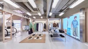 Bright, modern menswear retail interior with exposed concrete ceiling, linear fluorescent lighting, clothing racks, mannequins, and large digital screens displaying models.
