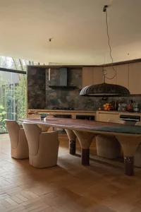 Modern dining room featuring a long curved walnut dining table with thick marble legs, beige upholstered chairs, a large woven pendant lamp, and a dramatic stone backsplash overlooking a bamboo garden.