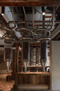 Industrial-style coffee roastery interior featuring three large stainless steel coffee bean hoppers hanging over a reclaimed wood counter with exposed beams and pipes.
