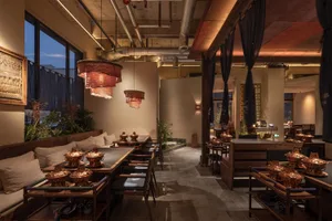 Modern industrial-style hotpot restaurant interior with long wooden communal tables, built-in cushioned banquettes, copper hotpots, and large red paper lanterns hanging from an exposed concrete and ductwork ceiling.