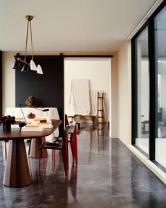 Modern minimalist dining room featuring a dark wood sculptural table, bright red dining chairs, polished concrete floor, black accent wall, and contemporary pendant light fixture.