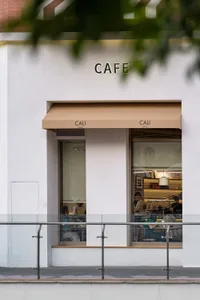 Street view of a modern white cafe named CALI with beige awnings, large glass windows showing customers inside, and a metal railing in the foreground.