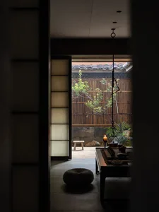 View from a dimly lit Japanese-style room through an open sliding door into a private bamboo courtyard garden with a low wooden table, woven cushion, and hanging iron lantern.