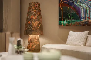 A tall table lamp with a large conical shade made of brown and orange Chinese floral brocade, lit and placed next to a cream-colored sofa with a white textured pillow, colorful abstract painting on the wall behind.