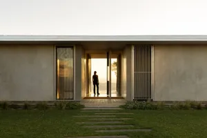 Modern minimalist concrete house with large glass doors and vertical metal screen, golden sunset light illuminating an open courtyard entrance with a woman standing on the wooden deck overlooking distant hills.