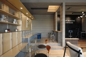 Modern cafe and livehouse interior featuring a long service counter with light birch plywood upper cabinets, blue glass mosaic tile wainscoting, round glass tables with molded plywood chairs, and a raised performance area in the background.